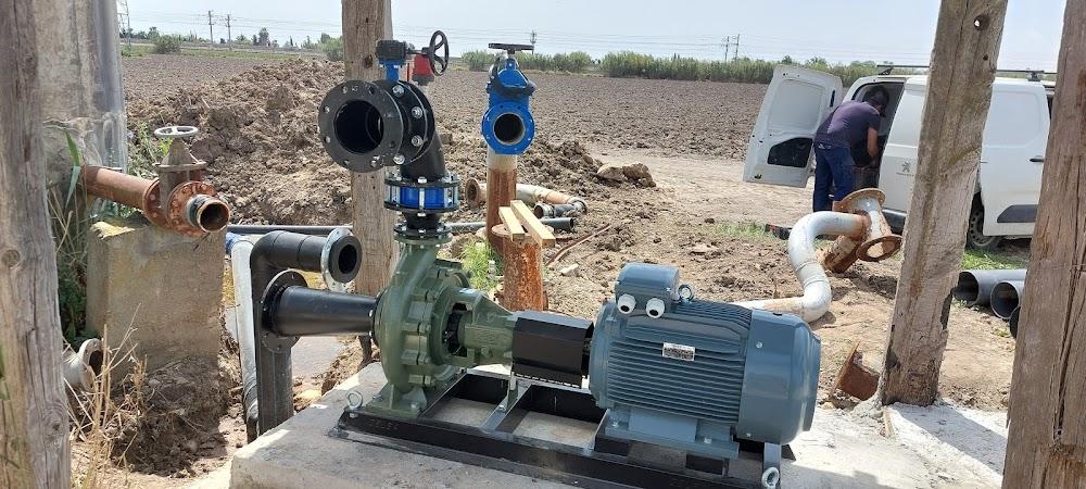 Una bomba de agua de gran tamaño con un motor eléctrico gris y cuerpo verde, conectada a tuberías y válvulas de color negro y azul. La instalación está sobre una base de concreto en un terreno de tierra abierto, y se puede ver una furgoneta blanca y una persona trabajando en segundo plano.