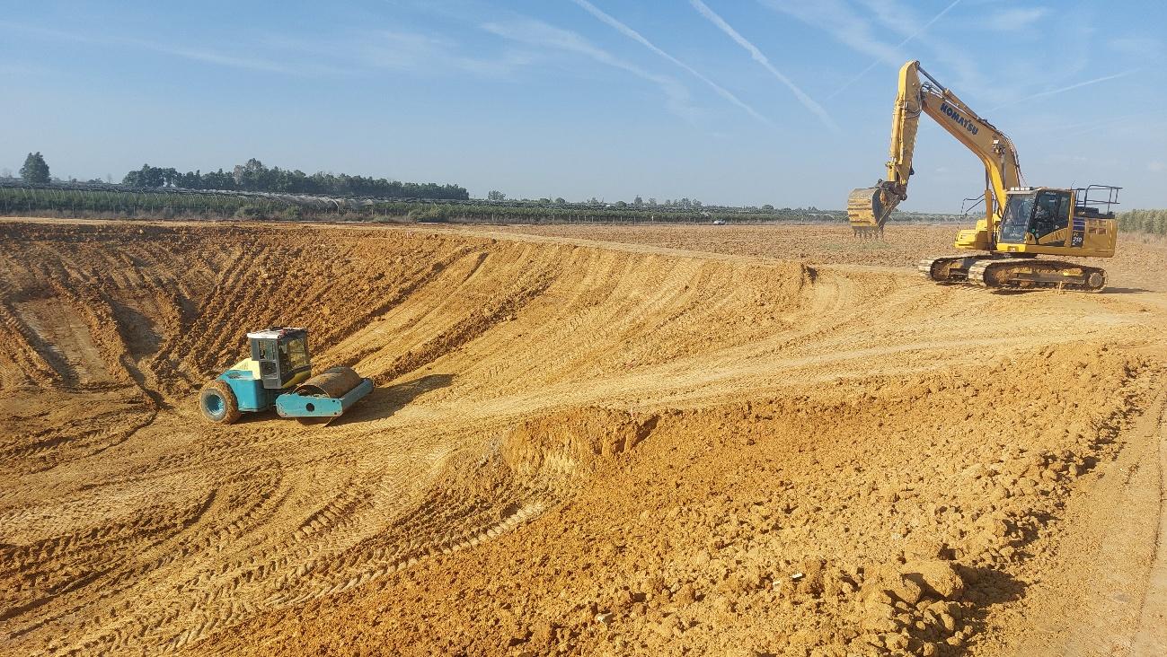 Excavadora amarilla y rodillo compactador azul trabajando en la construcción de una balsa o estanque en un terreno con tierra removida.