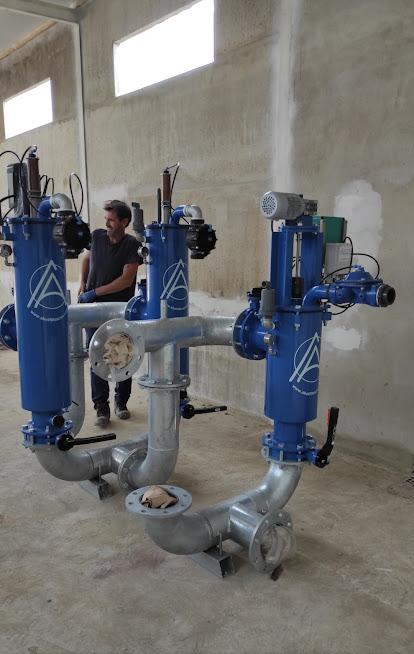 Sistema de filtración industrial de agua con tres unidades verticales azules y tuberías de gran diámetro, con un operario en segundo plano.