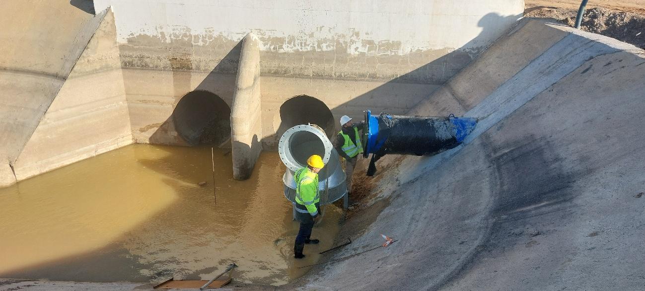 Dos trabajadores con equipo de protección instalando una tubería de gran diámetro en un canal de hormigón con agua.