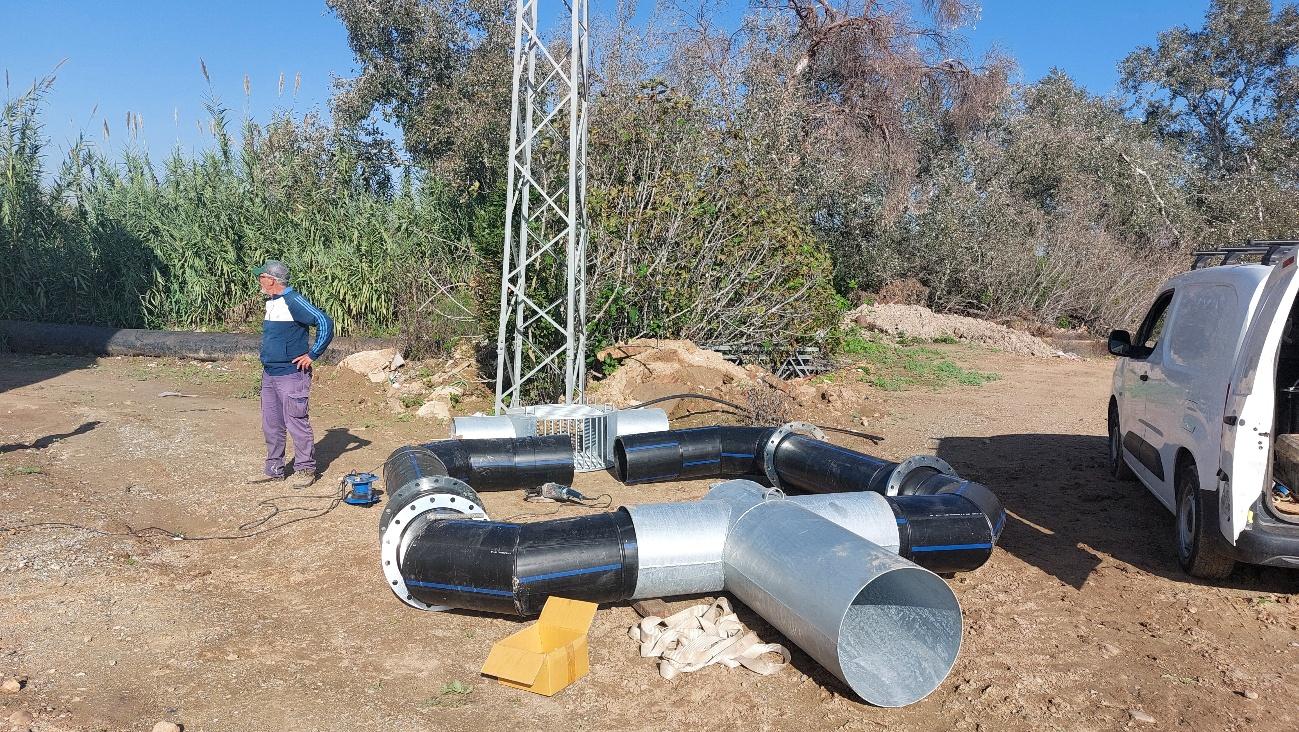 Grandes secciones de tubería con bridas y un trabajador supervisando su montaje en un terreno con una torre de metal y vegetación.