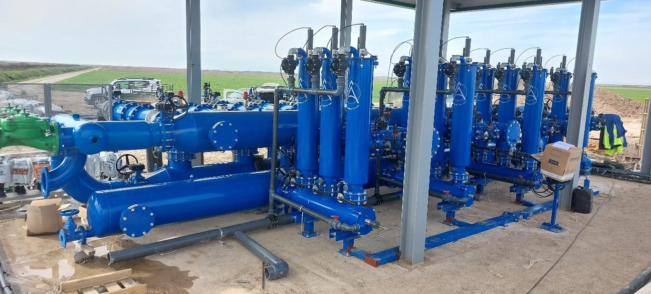 sistema-filtrado-riego-agricola Gran sistema de filtración de agua azul brillante y tuberías de riego en una instalación agrícola, con campos verdes al fondo.