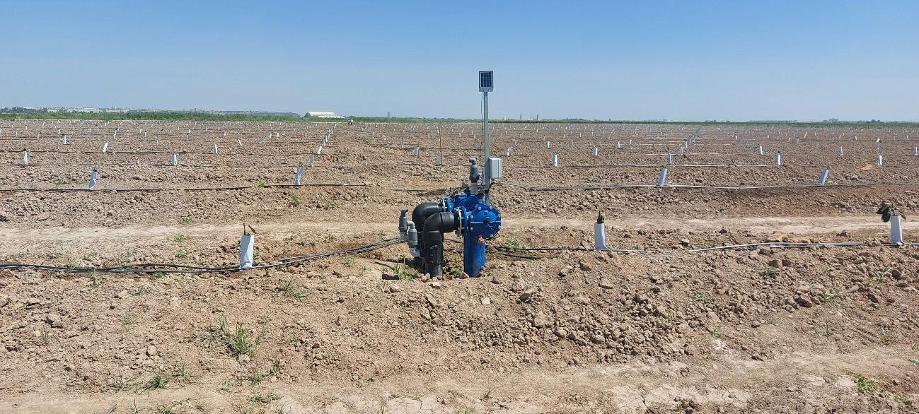 Sistema de riego con tuberías y válvulas azules, y un sensor solar, instalado en un campo de cultivo arado bajo un cielo despejado.