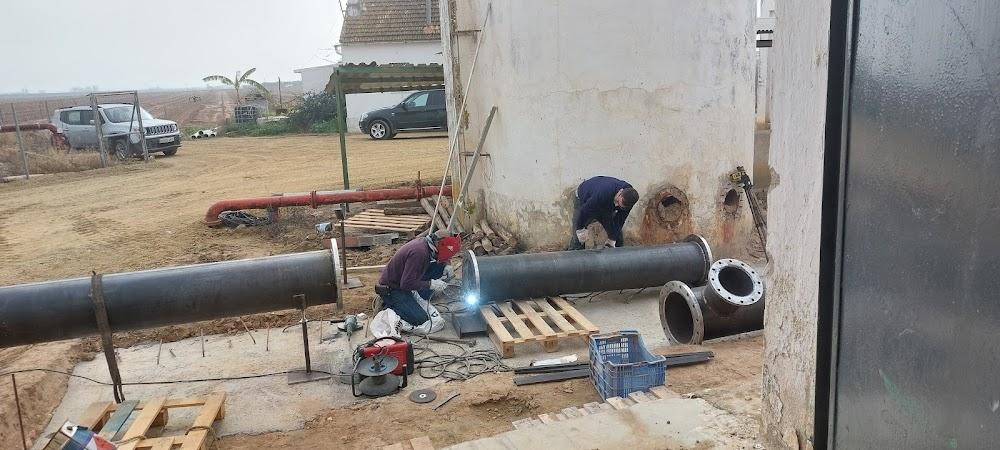 Dos trabajadores instalando y soldando grandes tuberías de metal en un sitio de construcción al aire libre, con un tanque de almacenamiento detrás.