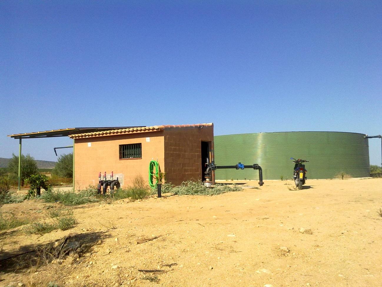 Gran tanque de almacenamiento de agua de color verde y una caseta de bombeo de ladrillo en un paisaje árido con cielo azul.